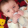 baby, bottle, milk, infant, dinosaur_print, onesie, blue_eyes, lying_down, blanket, red_blanket, yellow_text, hand, face, child, cute, indoors, closeup, feeding, young, person