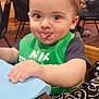 baby, child, tongue_out, green_bib, wooden_table, indoor, people, adult, window, chair, curly_hair, playful, cute, face, hand, high_chair, smiling, dining, family, room