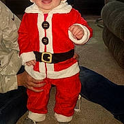 Skyler is registered to the contest to win money with this photo: toddler, child, santa_costume, red_clothing, white_fur_trim, indoor, carpet, smiling, holiday, festive, person, baby, sock, adult_hand, living_room, television, furniture, toy, happy, cute