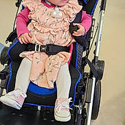 Athena is registered to the contest to win money with this photo: toddler, child, stroller, pink_dress, white_tights, shoes, hair_clips, indoor, floor, curious, seated, medical_device, baby_clothing, wheelchair, person, face, head, hands, footwear, expression