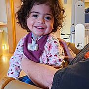 Athena is registered to the contest to win money with this photo: toddler, child, smiling, curly_hair, indoor, person, sitting, adult_arm, floral_clothing, chair, wooden_door, happy, home_interior, cozy, casual, portrait, baby, cute, warm_lighting, support