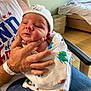 Gary is registered to the contest to win money with this photo: newborn, baby, infant, hat, blanket, hand, adult, hospital_wristband, chair, clothing, person, indoor, portrait, face, baby_hat, seated, holding, cute, wrapped, resting