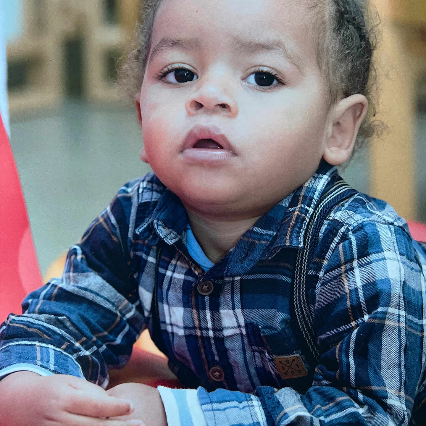 Djebril a rejoint le concours — aidez-le/la à gagner de superbes lots ! buttoned_shirt, casual_clothing, child, closeup, curious, expression, eyes, face, hair, hands, indoor, long_sleeves, person, plaid_shirt, portrait, red_surface, sitting, skin, toddler, young_child