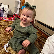 Maisie joined the competition — help win amazing prizes! toddler, child, smile, green_outfit, bow, carpet, indoor, toy, baby_wipes_box, heater, plastic_drawers, red_toy_car, floor, person, happy, candid, cute, sitting, home, clothing