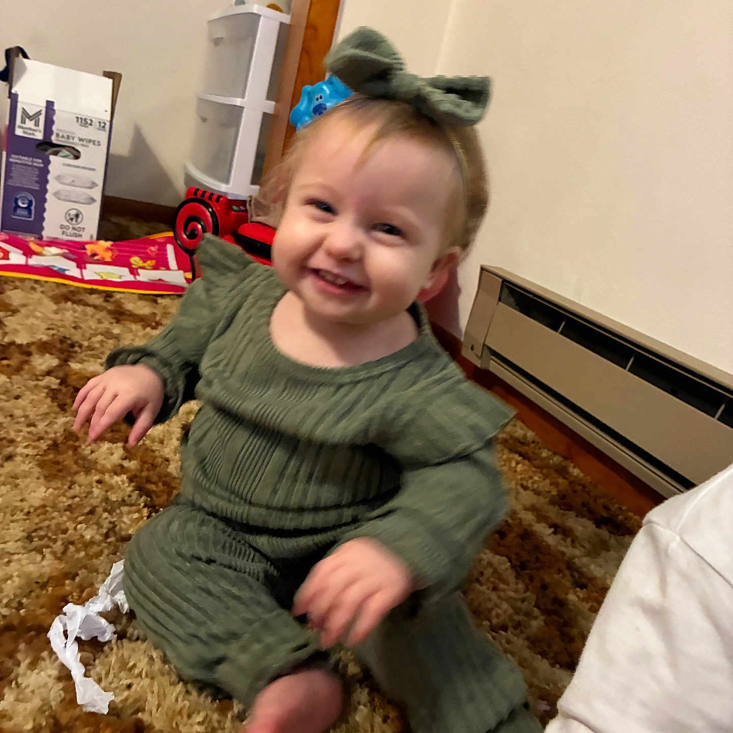 Maisie joined the competition — help win amazing prizes! toddler, child, smile, green_outfit, bow, carpet, indoor, toy, baby_wipes_box, heater, plastic_drawers, red_toy_car, floor, person, happy, candid, cute, sitting, home, clothing