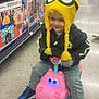 Robert is registered to the contest to win money with this photo: aisle, black_jacket, braids, child, colorful_boxes, cute, floor, gray_pants, happy, hippo_toy, indoor, kid, minion_hat, pink_toy, playful, smiling, sneakers, toy_ride, toy_store, yellow_hat