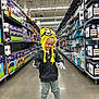 balloons, boxes, child, concrete_floor, gloves, hoodie, indoor, kid, minion_hat, pants, party_supplies, person, playful, retail, shelves, shopping, smiling, sneakers, store_aisle, yellow_hat