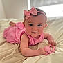 baby, child, pink_bow, headband, pink_dress, toy, heart_shaped_toy, blanket, bed, infant, cute, smiling, portrait, indoors, soft_light, close_up, person, skin, face, happy