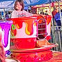 Bryce is registered to the contest to win money with this photo: child, teacup_ride, carnival, amusement_park, colorful, ride, fair, young_person, outdoor, fun, entertainment, crowd, background, food_stand, purple, red, yellow, hair, clothing, person
