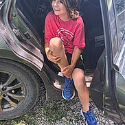 Bryce is registered to the contest to win money with this photo: child, car, open_door, red_tshirt, blue_sneakers, gravel, grass, smiling, casual, outdoor, sunlight, shadow, shorts, sitting, person, hair, hand, footwear, vehicle, relaxed