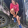 child, car, open_door, red_tshirt, blue_sneakers, gravel, grass, smiling, casual, outdoor, sunlight, shadow, shorts, sitting, person, hair, hand, footwear, vehicle, relaxed