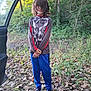 child, forest, outdoor, leaves, trees, nature, person, standing, clothing, long_hair, shoes, car_door, casual, blue_pants, patterned_shirt, greenery, daylight, young, smile, adventure