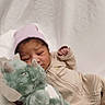 baby, infant, child, sleepy, portrait, striped_outfit, knit_hat, pink_hat, plush_toy, dinosaur_toy, stuffed_animal, bed, bedding, indoor, cozy, lying_down, raised_fist, soft_lighting, white_background, cute