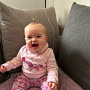 Maëly a rejoint le concours — aidez-le/la à gagner de superbes lots ! baby, child, smiling, sitting, pink_clothing, floral_pants, pony_tail, cushion, blanket, indoor, happy, cute, portrait, young_child, cozy, furniture, person, home, infant, expression