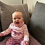 baby, child, smiling, sitting, pink_clothing, floral_pants, pony_tail, cushion, blanket, indoor, happy, cute, portrait, young_child, cozy, furniture, person, home, infant, expression