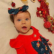 Sofia a rejoint le concours — aidez-le/la à gagner de superbes lots ! baby, child, face, blue_eyes, red_dress, bow, christmas_ornaments, holiday_decor, lying_down, cute, surprised_expression, infant, festive, indoor, celebration, toy, decoration, person, smiling, headband