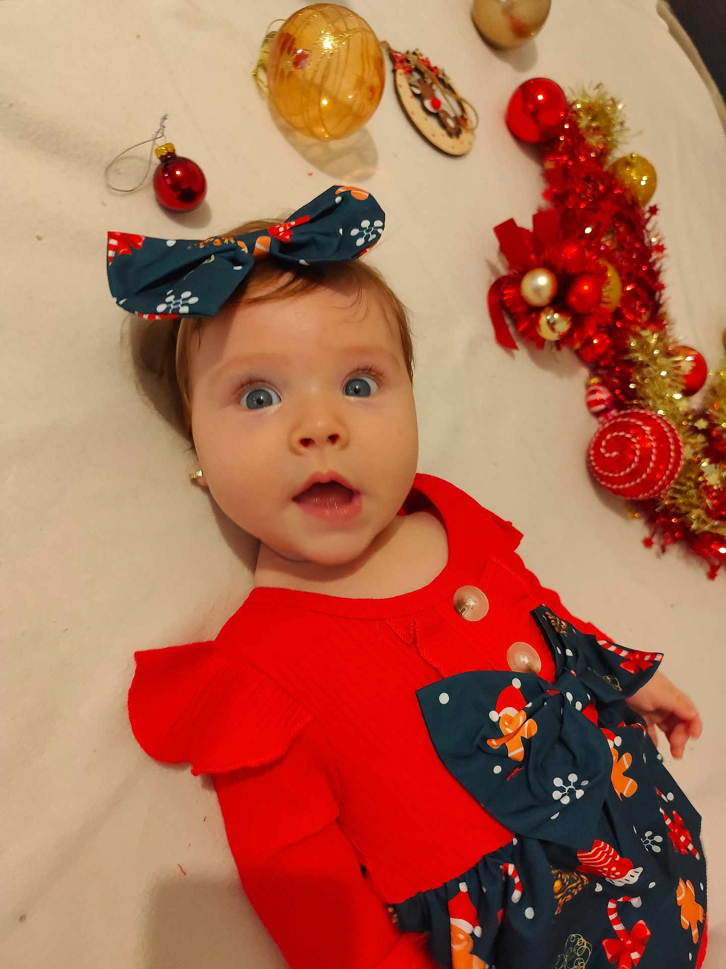 Sofia a rejoint le concours — aidez-le/la à gagner de superbes lots ! baby, child, face, blue_eyes, red_dress, bow, christmas_ornaments, holiday_decor, lying_down, cute, surprised_expression, infant, festive, indoor, celebration, toy, decoration, person, smiling, headband