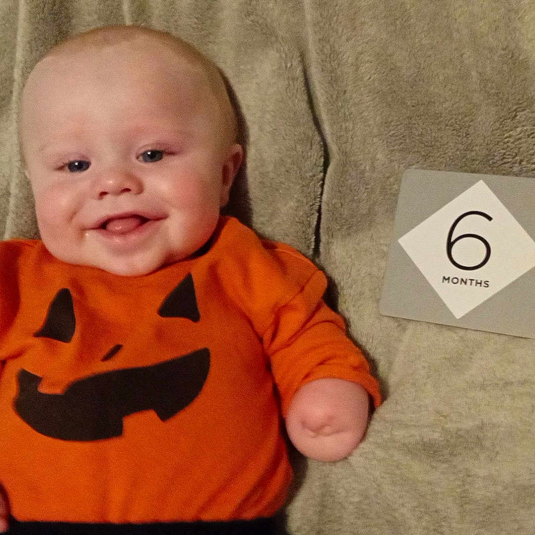 Christian is registered to the contest to win money with this photo: 6_months, arm, baby, blanket, child, cute, face, happy, head, indoors, infant, lying_down, milestone_card, orange_clothing, person, pumpkin_face, skin, smile, soft_texture, wearing
