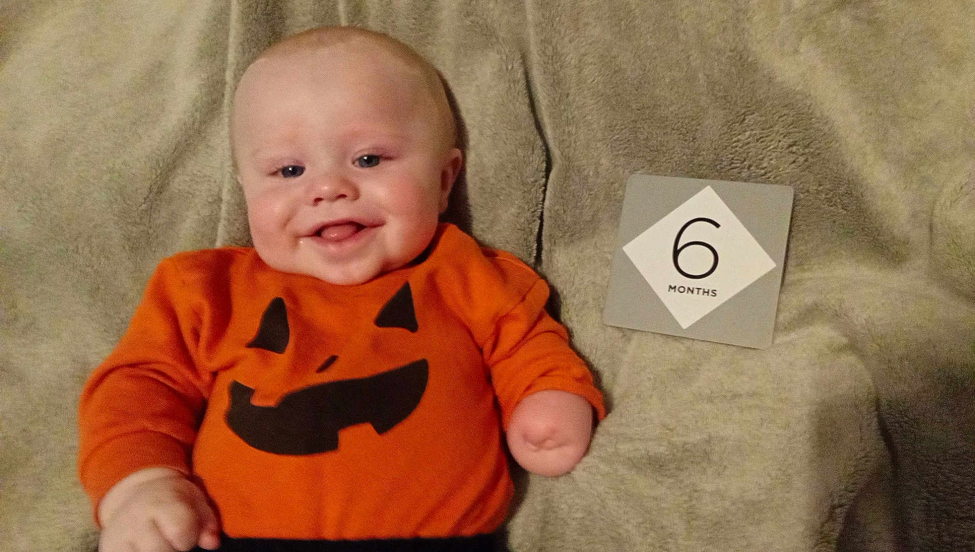 Christian is registered to the contest to win money with this photo: baby, infant, orange_clothing, pumpkin_face, milestone_card, 6_months, blanket, smile, happy, cute, lying_down, child, face, head, skin, arm, person, indoors, soft_texture, wearing