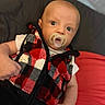 Ledger is registered to the contest to win money with this photo: baby, pacifier, vest, plaid, red, black, white, infant, face, eyes, hand, clothing, lying_down, indoor, soft_surface, portrait, cute, child, young, person