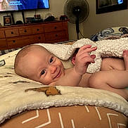 Carson is registered to the contest to win money with this photo: baby, blanket, bed, smile, face, infant, indoor, cozy, furniture, dresser, fan, diaper, skin, hand, foot, room, portrait, lying_down, comfort, cute