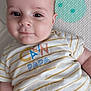 baby, child, face, smile, striped_shirt, text_on_clothing, lying_down, patterned_background, cute, infant, person, young_child, closeup, head, skin, eyes, cheeks, short_sleeves, casual_clothing, indoor