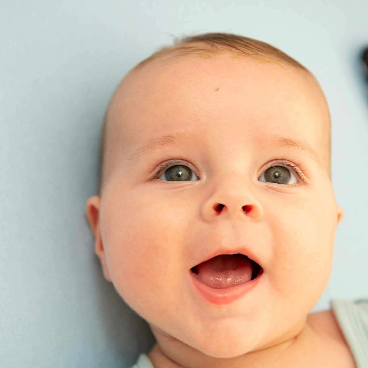 Raphaël participe au concours pour gagner de l'argent avec cette photo : baby, baby_clothing, blue_background, cheerful, child, close_up, cute, expression, eyes, face, happy, head, indoors, infant, mouth_open, patterned_cushion, portrait, skin, smile, young
