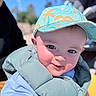 baby, child, smiling, cap, hat, blue_sky, outdoor, vest, puffy_jacket, close_up, face, happy, sunlight, cute, person, portrait, daylight, casual_clothing, headwear, infant