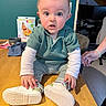 baby, child, table, green_sweater, striped_pants, white_shoes, indoor, person, wide_eyes, curious, hand, wood, floor, box, background, clothing, sitting, expression, face, home