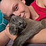 affection, animal, blue_eyes, cat, close_up, cozy, cuddling, cute, geometric_pattern, gray_cat, green_eyes, human_animal_bond, indoor, person, pet, pillow, pink_tank_top, relaxed, resting, shaved_head
