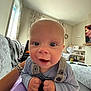 Oliver is registered to the contest to win money with this photo: baby, infant, face, smile, hands, clothing, overalls, living_room, couch, blanket, window, curtains, fan, television, remote_control, shelf, decor, person, indoor, cute