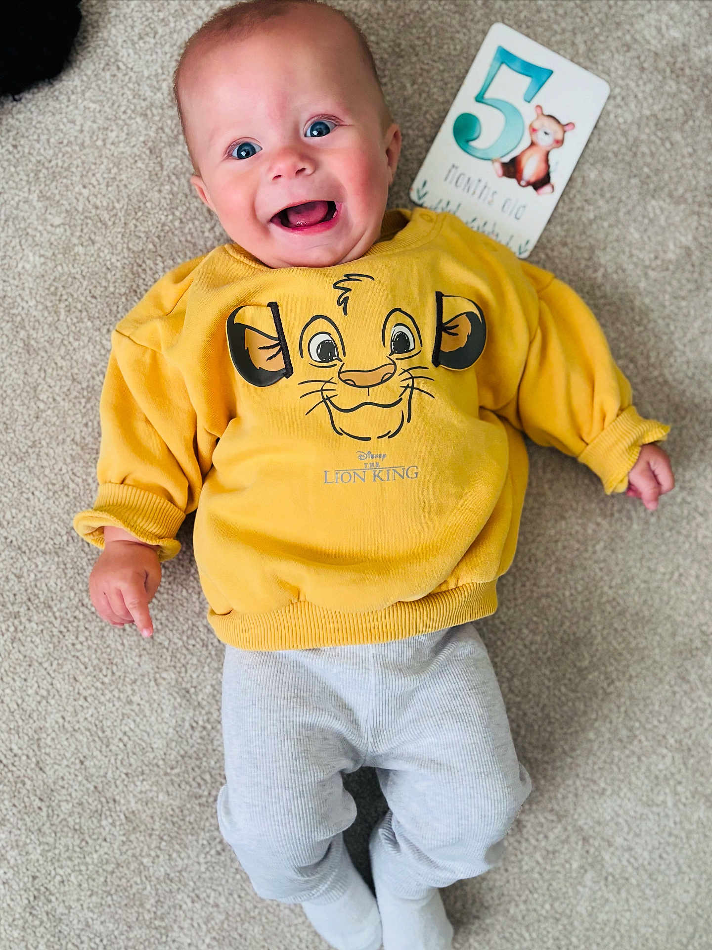 Oliver is registered to the contest to win money with this photo: baby, infant, smiling, yellow_sweatshirt, lion_king, milestone_card, 5_months_old, blue_eyes, carpet, laying_down, cute, happy, child, clothing, sleeves, pants, feet, portrait, indoor, person