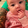 baby, infant, child, portrait, close_up, sitting, curious, finger_in_mouth, pink_stripes, onesie, indoors, adult, tattoo, family, candid, face, eyes, hands, couch, cozy