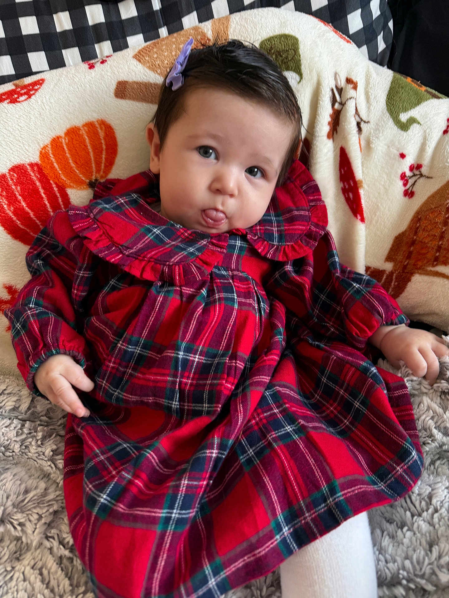 Noelle is registered to the contest to win money with this photo: baby, child, plaid_dress, red_clothing, blanket, autumn_theme, pumpkin_pattern, cozy, cute, tongue_out, hair_clip, indoor, seated, smiling, infant, soft_texture, person, portrait, young_child, warm_colors