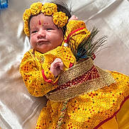 Vansh joined the competition — help win amazing prizes! baby, child, yellow_clothing, floral_headband, peacock_feather, traditional_dress, infant, portrait, face, smile, lying_down, fabric_background, cute, hand, decorative_belt, indoors, closeup, colorful, soft_light, celebration