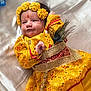 baby, child, yellow_clothing, floral_headband, peacock_feather, traditional_dress, infant, portrait, face, smile, lying_down, fabric_background, cute, hand, decorative_belt, indoors, closeup, colorful, soft_light, celebration