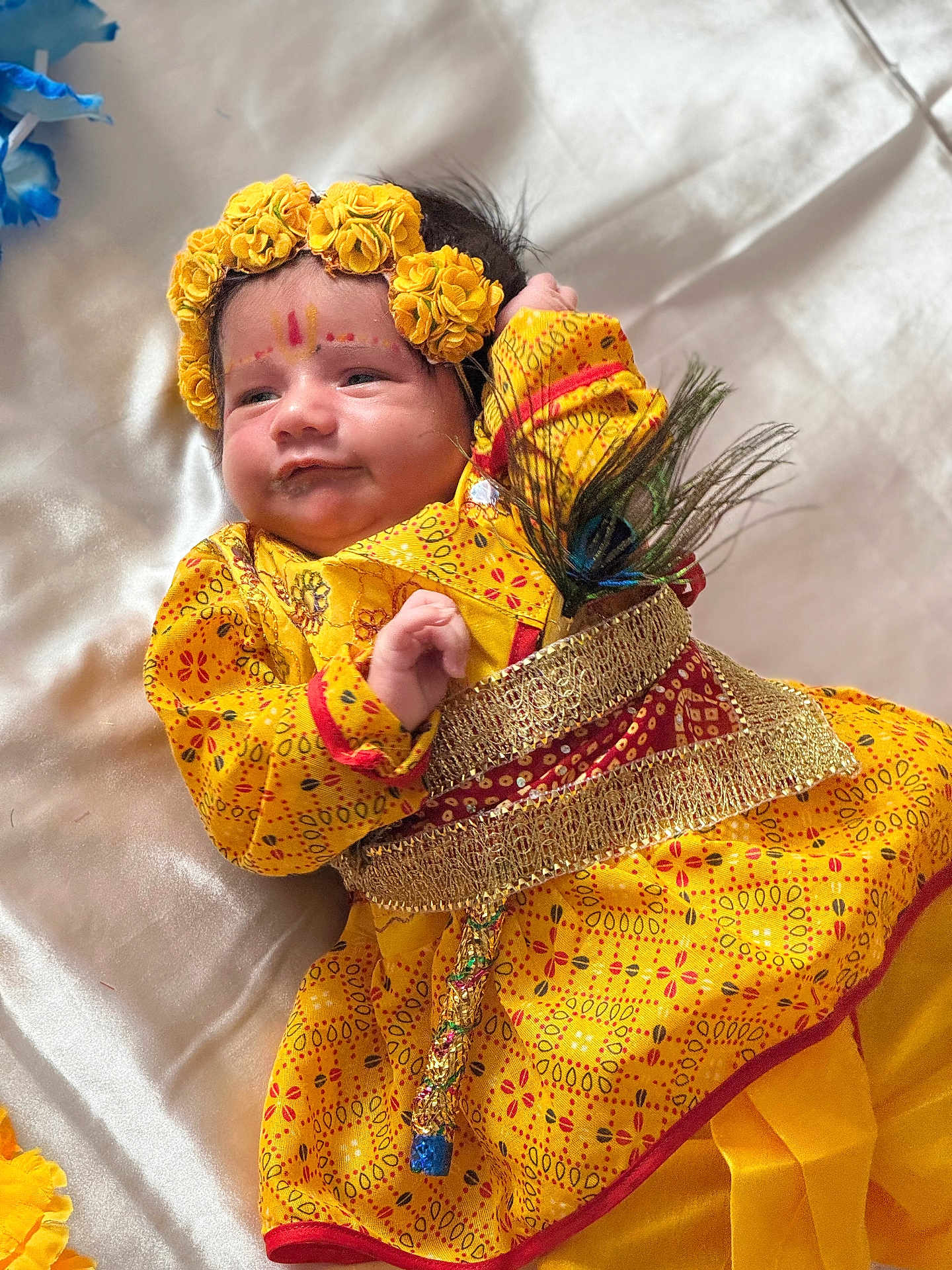 Vansh joined the competition — help win amazing prizes! baby, child, yellow_clothing, floral_headband, peacock_feather, traditional_dress, infant, portrait, face, smile, lying_down, fabric_background, cute, hand, decorative_belt, indoors, closeup, colorful, soft_light, celebration