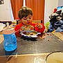 child, toddler, bib, food, messy_face, bowl, plate, sippy_cup, table, chair, spoon, curtains, living_room, meal, eating, food_spill, toys, clothing, highchair, timestamp