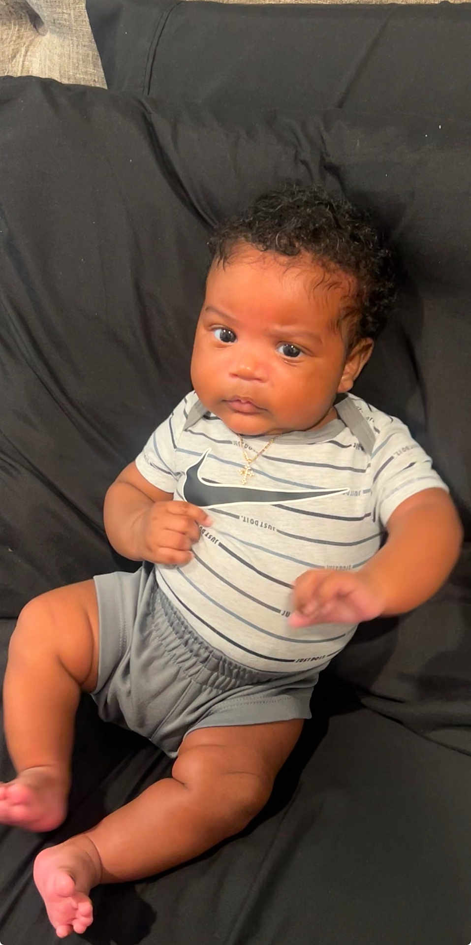 Zaylan is registered to the contest to win money with this photo: baby, infant, child, curly_hair, striped_shirt, nike_logo, shorts, gold_necklace, cross_pendant, couch, black_cushion, skin, legs, feet, hands, face, eyes, expression, indoors, portrait