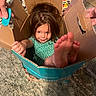 child, cardboard_box, playful, indoor, carpet, toy, hands, feet, smiling, girl, green_dress, hair, cozy, fun, family, home, cute, young, person, relaxed