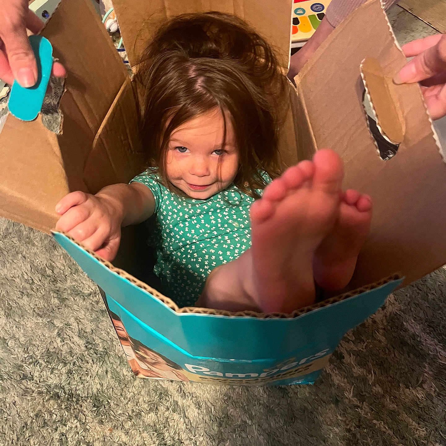 Mötley joined the competition — help win amazing prizes! child, cardboard_box, playful, indoor, carpet, toy, hands, feet, smiling, girl, green_dress, hair, cozy, fun, family, home, cute, young, person, relaxed