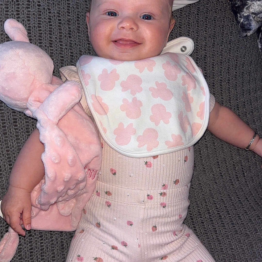 Romi is registered to the contest to win money with this photo: baby, bib, child, clothing, cute, fabric, face, happy, indoors, infant, lying_down, patterned_clothing, person, pink, plush_toy, portrait, smiling, socks, soft_toy, texture