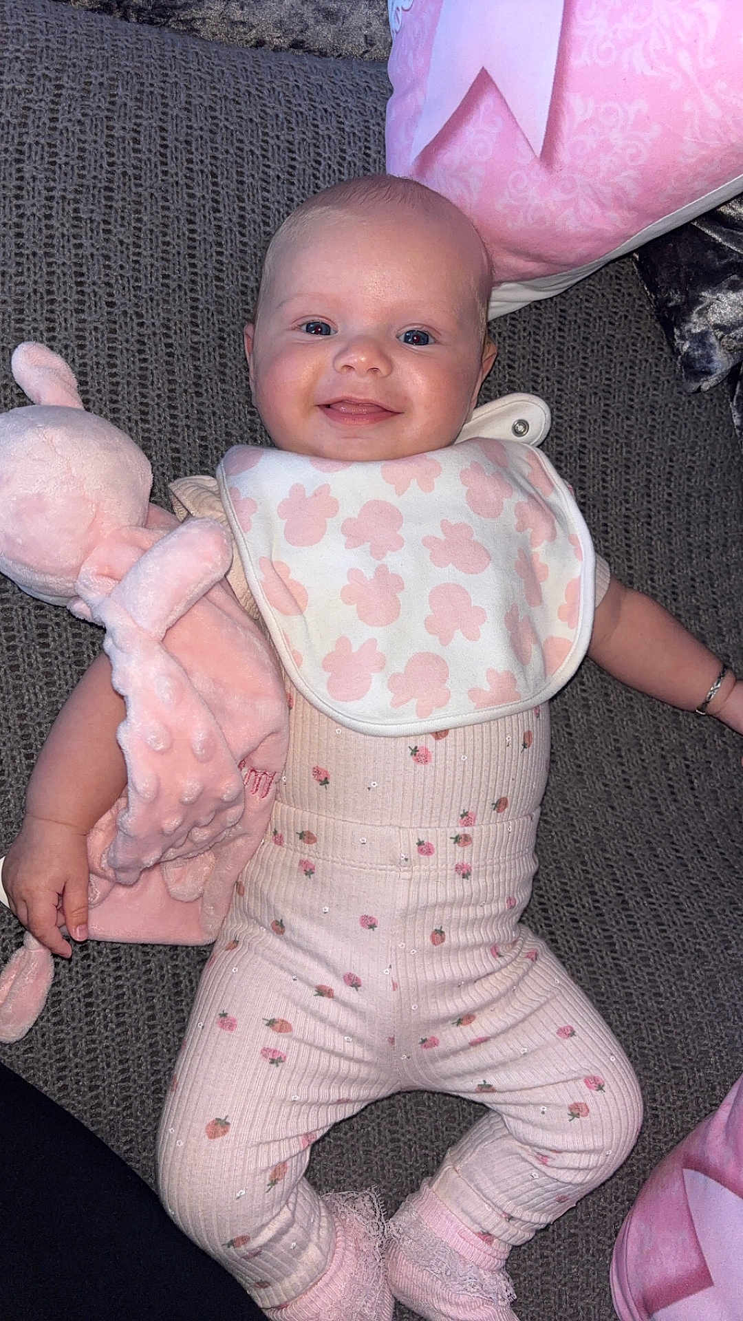 Romi is registered to the contest to win money with this photo: baby, infant, smiling, bib, plush_toy, pink, clothing, lying_down, happy, face, person, cute, soft_toy, socks, patterned_clothing, indoors, texture, fabric, child, portrait