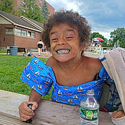 Ja'Kobe Anthony is registered to the contest to win money with this photo: child, smiling, blue_floatation_device, sailboat_pattern, water_bottle, picnic_table, outdoor, grass, brick_building, pool_area, people, cloudy_sky, curly_hair, happy, summer, playful, water, umbrella, towel, hand