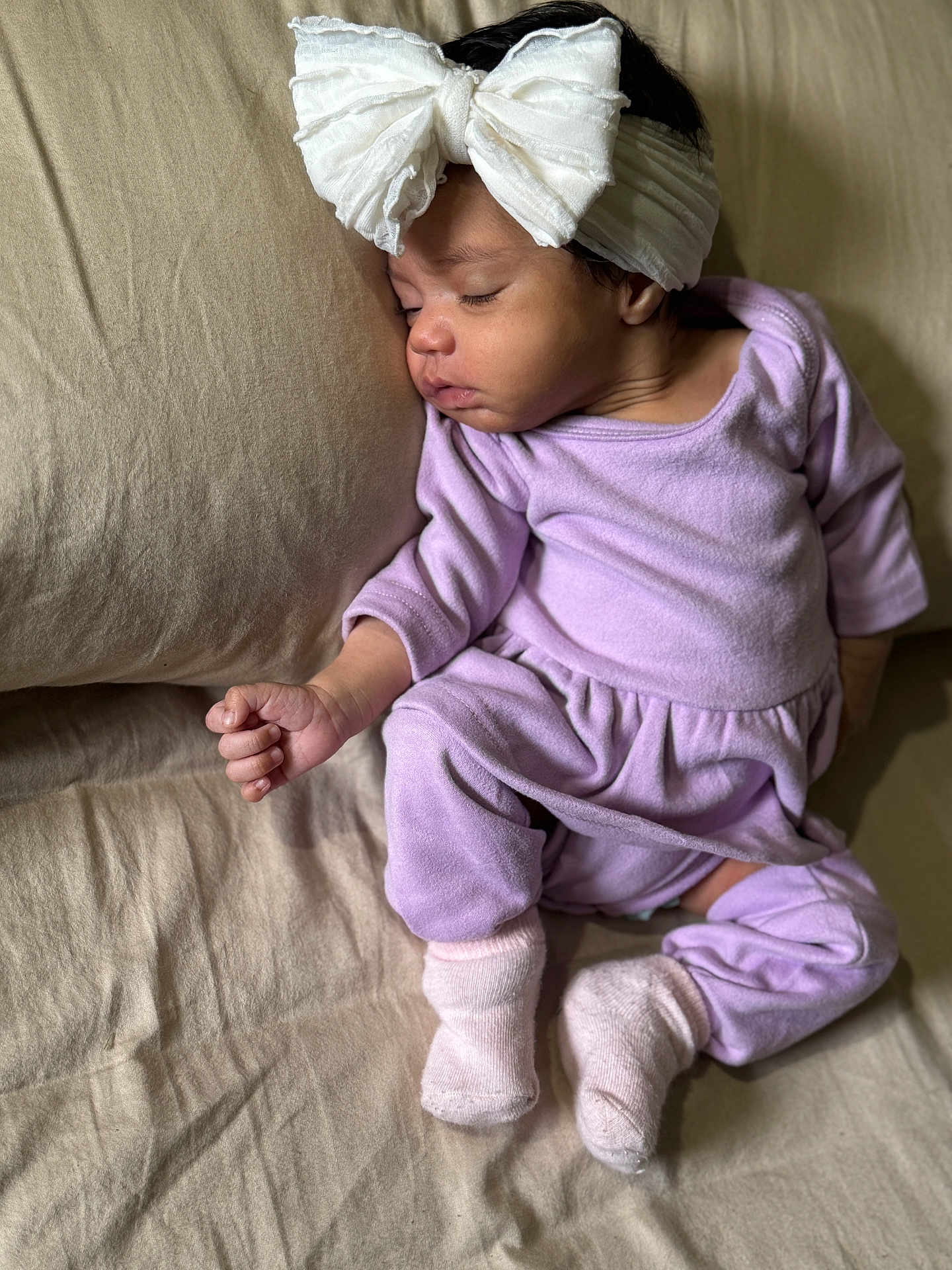 Adaly is registered to the contest to win money with this photo: baby, sleeping, headband, bow, purple_outfit, socks, couch, pillow, bed, infant, child, rest, soft_fabric, indoors, peaceful, skin, hand, footwear, clothing, portrait