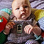 Ezra is registered to the contest to win money with this photo: baby, infant, child, overalls, plush_toy, cushion, colorful, indoor, portrait, cute, clothing, face, hands, sitting, expression, soft, comfort, pacifier, pattern, close_up