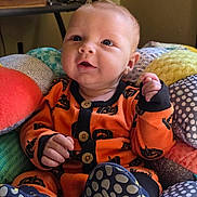 Ezra is registered to the contest to win money with this photo: baby, infant, child, orange_clothing, pumpkin_pattern, cushion, colorful, indoor, seated, cute, happy, blue_eyes, hand, footwear, soft_toys, fabric, portrait, smile, cozy, home