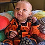 baby, infant, child, orange_clothing, pumpkin_pattern, cushion, colorful, indoor, seated, cute, happy, blue_eyes, hand, footwear, soft_toys, fabric, portrait, smile, cozy, home