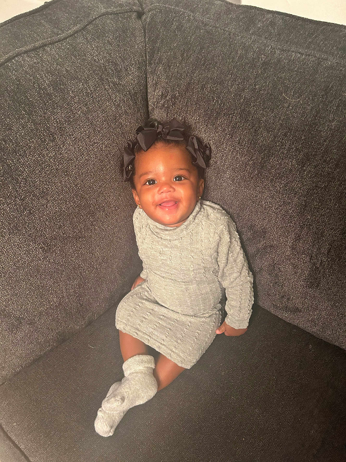 Nevaeh joined the competition — help win amazing prizes! toddler, child, smile, sitting, couch, gray_dress, knitted_clothing, socks, hair_bows, indoor, happy, portrait, baby, cute, cozy, furniture, person, face, skin, head