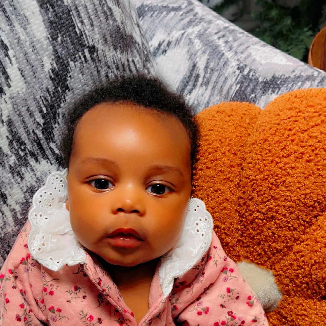 Janiyah is registered to the contest to win money with this photo: baby, infant, child, pink_clothing, floral_pattern, white_collar, couch, pillow, orange_pillow, indoor, portrait, face, curly_hair, cute, seated, looking_at_camera, soft_lighting, home, cozy, young_child
